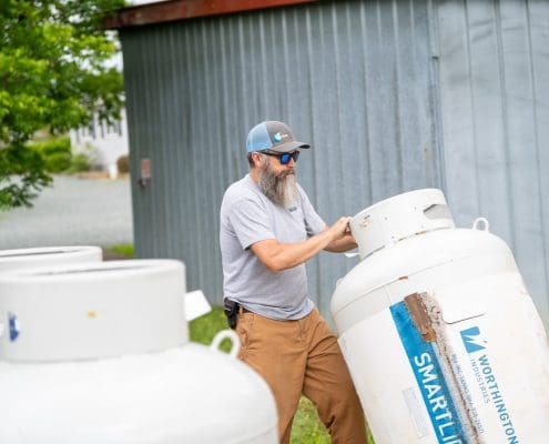 A man in a gray shirt and cap inspects a propane tank outdoors, near a metal building