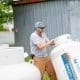 A man in a gray shirt and cap inspects a propane tank outdoors, near a metal building